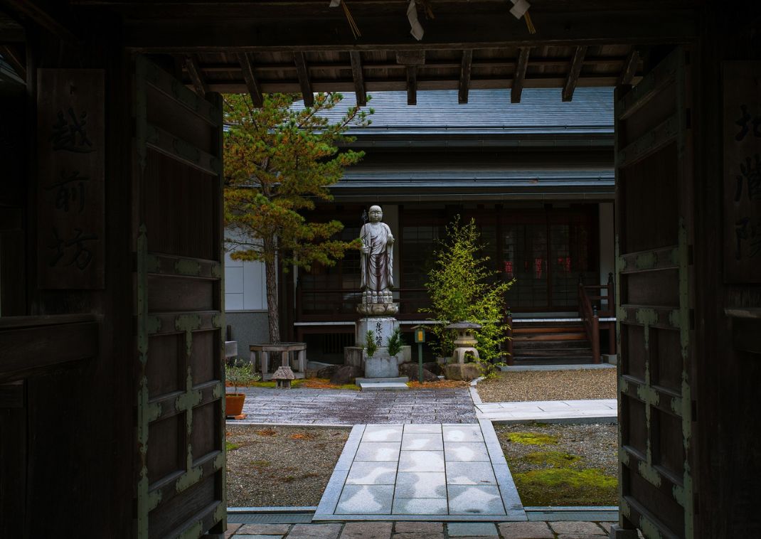 Indgang til en traditionel japansk gårdhave med statue, træer og stenlanterne foran et træhus