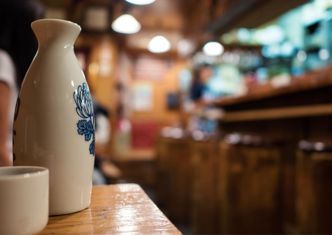 Sakeflaske og kop med restaurant ude af fokus i baggrunden.