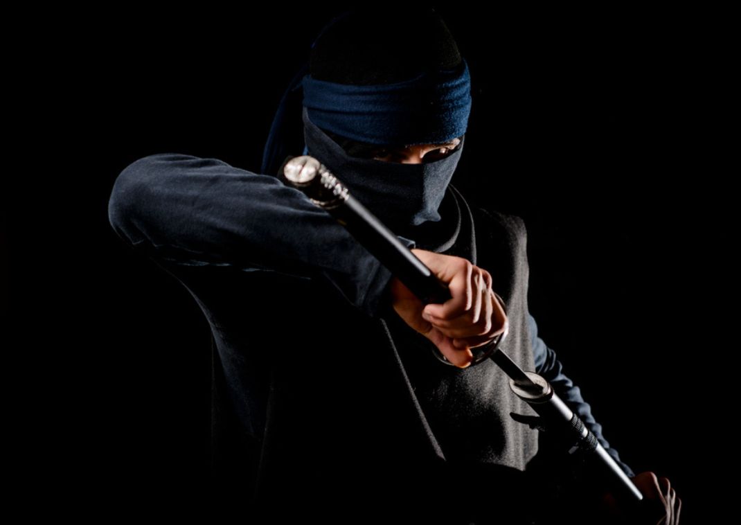 En ninja klædt i sort tøj og ansigtsmaske holder et sværd i en forsvarsposition mod en mørk baggrund.