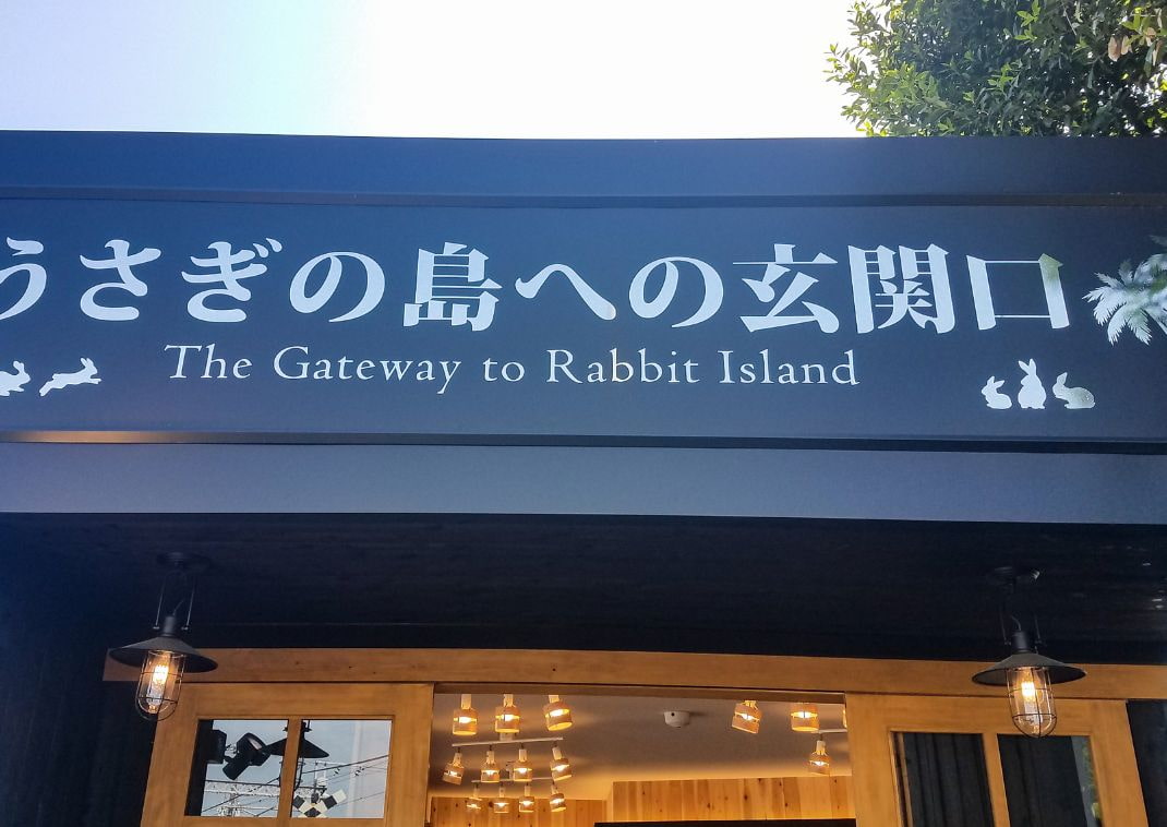 Indgangen til en butik med et skilt, der lyder -The Gateway to Rabbit Island- på både japansk og engelsk, dekoreret med kaninsilhuetter og oplyst af hængende lamper.